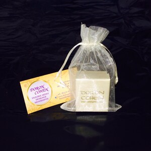 May include: A small, white box with gold lettering that reads "Doron Cohen" and "www.doroncohen.com". The box is sitting on a white, sheer fabric bag. A business card with the same name and the text "Filigree Artist Designer Silversmith" is visible in the background.