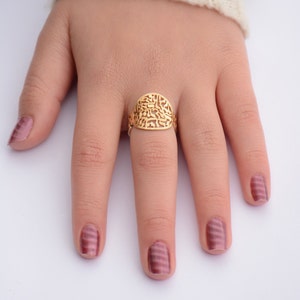 May include: A gold ring with a filigree design featuring Hebrew letters. The ring is worn on the finger of a hand with pink nail polish.