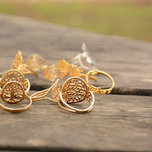May include: A collection of gold-toned rings with intricate designs, including a tree of life motif, on a wooden surface.