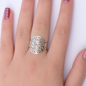 K&ouml;nnte beinhalten: Ein silberner Ring mit einem runden, durchbrochenen Design mit hebr&auml;ischen Buchstaben. Der Ring wird an einem Finger mit rosafarbenem Nagellack getragen.