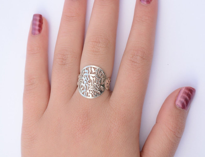 K&ouml;nnte beinhalten: Ein silberner Ring mit einem runden, durchbrochenen Design mit hebr&auml;ischen Buchstaben. Der Ring wird an einem Finger mit rosafarbenem Nagellack getragen.