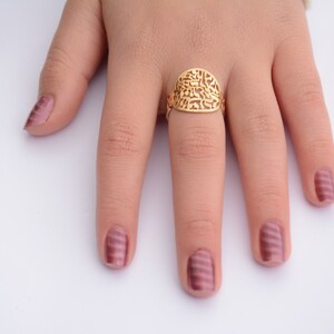 May include: A gold ring with a filigree design featuring Hebrew lettering. The ring is worn on the middle finger of a hand with pink nail polish.