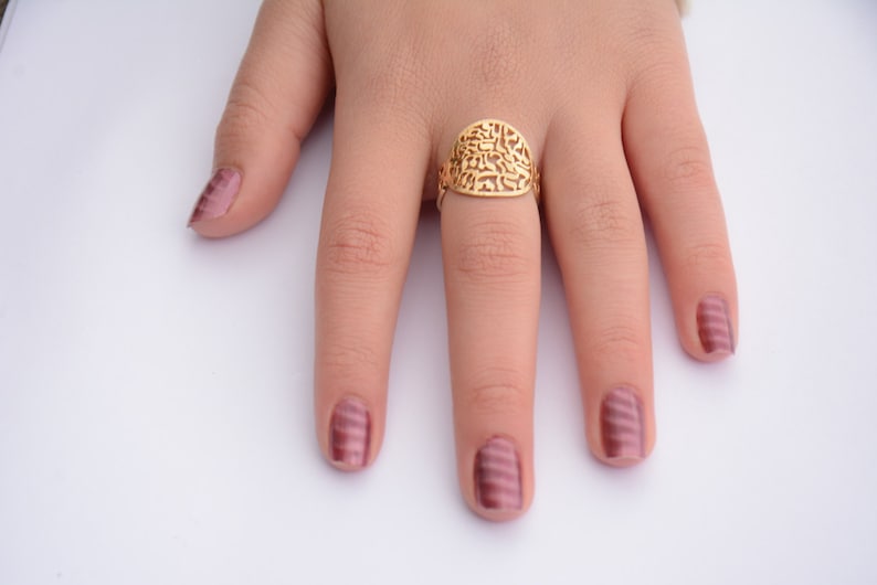 May include: A gold ring with a filigree design featuring Hebrew lettering. The ring is worn on the middle finger of a hand with pink nail polish.