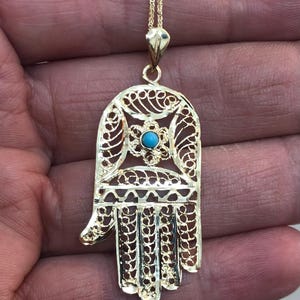Könnte beinhalten: Vergoldete Hamsa-Hand-Anhänger-Halskette. Der Anhänger hat ein filigranes Design mit einem türkis farbenen Stein in der Mitte. Die Halskette hat eine zarte Kette und ist ein Symbol für Schutz und Glück.