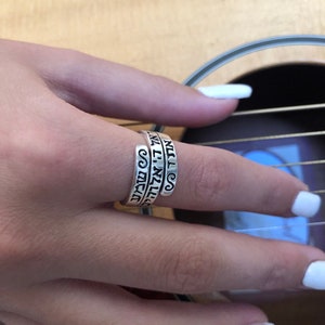 May include: A silver ring with the words "ALL I NEED" and "YOU" engraved on it. The ring is worn on a finger.