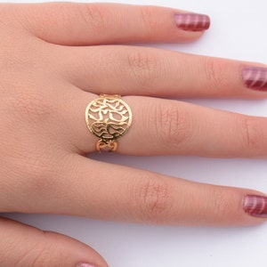 May include: A gold ring with a circular design featuring intricate cutouts. The ring is worn on the middle finger of a hand with pink nail polish.