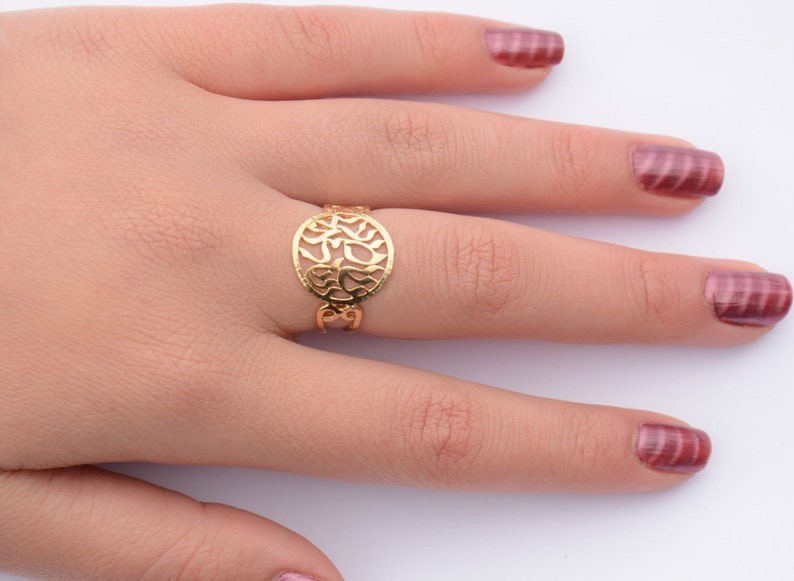 May include: A gold ring with a circular design featuring intricate cutouts. The ring is worn on the middle finger of a hand with pink nail polish.