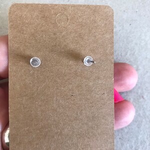 May include: A pair of silver stud earrings with clear backs, shown on a brown paper card.