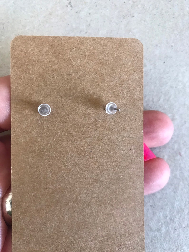 May include: A pair of silver stud earrings with clear backs, shown on a brown paper card.