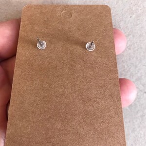 May include: Close-up of the back of a pair of stud earrings attached to a brown cardboard earring display card. The earrings have silver posts and clear plastic backs. The card has a small hole at the top for hanging.