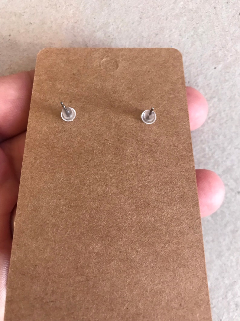 May include: Close-up of the back of a pair of stud earrings attached to a brown cardboard earring display card. The earrings have silver posts and clear plastic backs. The card has a small hole at the top for hanging.
