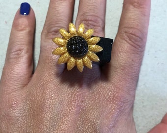 Könnte beinhalten: Ein goldener Sonnenblumenring mit schwarzem Zentrum, auf einem schwarzen quadratischen Band. Der Ring wird an einer Hand mit blauem Nagellack getragen.