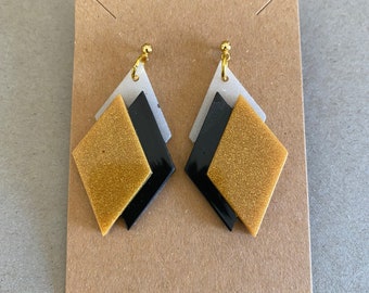 Könnte beinhalten: Ein Paar geometrische Ohrringe in Gold, Schwarz und Silber. Die Ohrringe sind diamantförmig und übereinander gelegt.