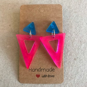 May include: A pair of handmade earrings featuring a neon pink triangle design with a blue triangle accent. The earrings are attached to a brown card with the text "Handmade with love".