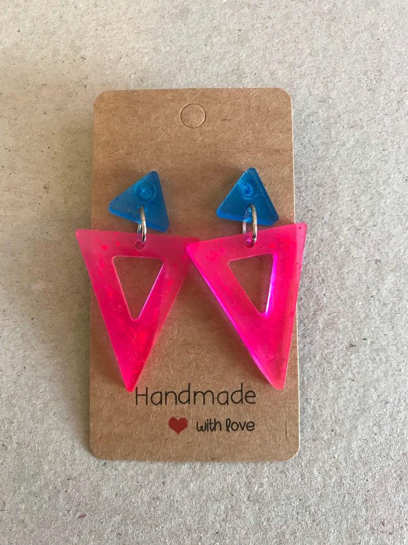 May include: A pair of handmade earrings featuring a neon pink triangle design with a blue triangle accent. The earrings are attached to a brown card with the text "Handmade with love".