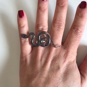 May include: A silver snake ring with glitter embedded in the resin. The ring is worn on the middle finger of a hand with red nail polish.
