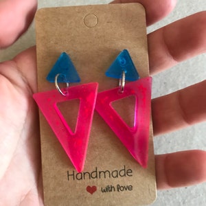 May include: A pair of handmade earrings featuring a neon pink triangle with a smaller blue triangle on top. The earrings are attached to a brown card that reads "Handmade with love."