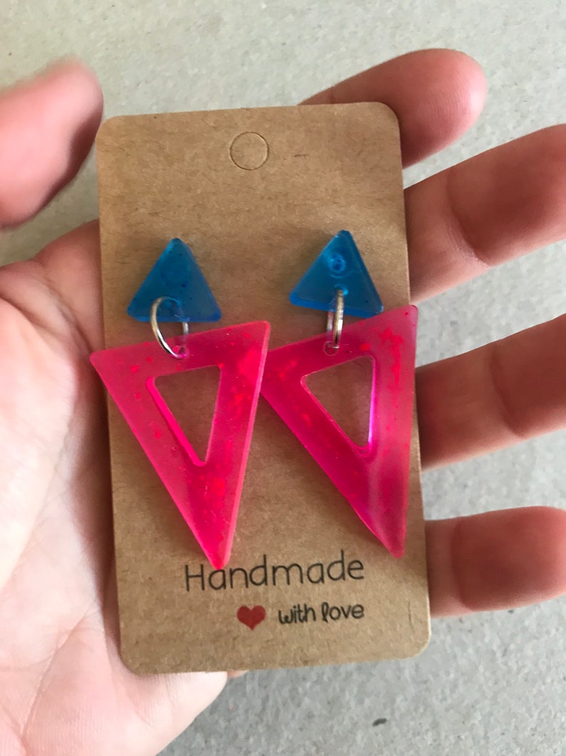 May include: A pair of handmade earrings featuring a neon pink triangle with a smaller blue triangle on top. The earrings are attached to a brown card that reads "Handmade with love."