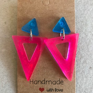 May include: A pair of handmade earrings featuring a blue triangle stud and a larger, open pink triangle. The earrings are attached to a brown card with the text "Handmade with love".
