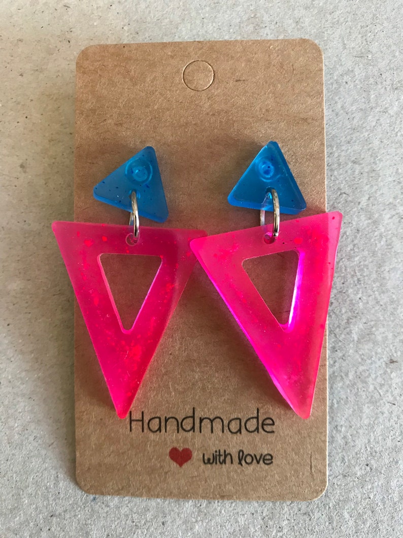 May include: A pair of handmade earrings featuring a blue triangle stud and a larger, open pink triangle. The earrings are attached to a brown card with the text "Handmade with love".