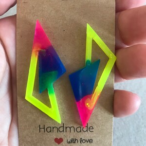 Puede incluir: Un par de pendientes hechos a mano con un contorno amarillo ne&oacute;n y un interior de colores arco&iacute;ris. Los pendientes tienen forma de rayo y se muestran en una tarjeta marr&oacute;n con el texto "Handmade with love".