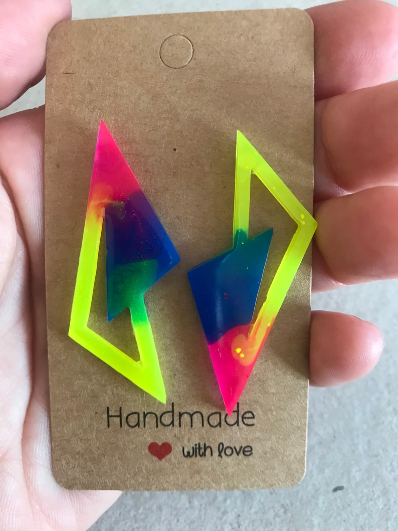 Puede incluir: Un par de pendientes hechos a mano con un contorno amarillo ne&oacute;n y un interior de colores arco&iacute;ris. Los pendientes tienen forma de rayo y se muestran en una tarjeta marr&oacute;n con el texto "Handmade with love".