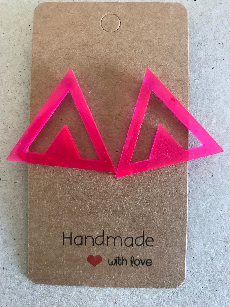 May include: A pair of pink triangle shaped earrings with a cutout design. The earrings are on a brown card with the text "Handmade with love".