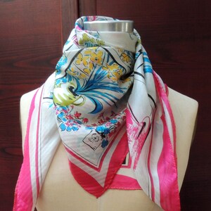 May include: A white silk scarf with a pink border and a colorful floral print. The scarf is tied around a mannequin's neck.