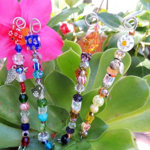 May include: Four beaded garden stakes with wire loops for hanging. Each stake features a unique design with colorful glass beads, charms, and crystals. The stakes are set against a backdrop of green foliage and a pink flower.