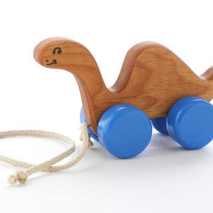 May include: A wooden toy dinosaur with blue wheels and a pull string. The dinosaur has a painted face with a smile.