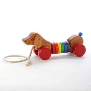 May include: Wooden pull-along toy dog with a smiling face, ears, and a blue ball tail. The dog's body is made of colorful rainbow rings and red wheels. A beige rope is attached for pulling.