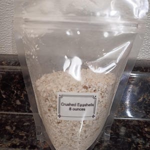 Könnte beinhalten: Ein klarer Plastikbeutel gefüllt mit zerstoßenen Eierschalen. Ein weißes Etikett mit schwarzem Text lautet "Crushed Eggshells 8 ounces". Die Eierschalen sind hellbeige. Der Beutel ist oben versiegelt.