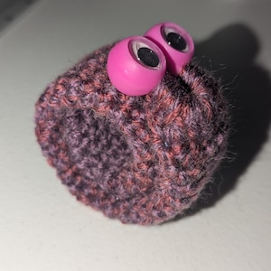 May include: A small knitted hat with pink googly eyes. The hat is a dark brown and pink color.