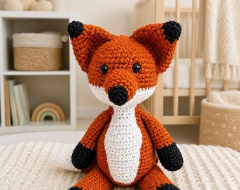 May include: A crochet fox stuffed animal featuring a burnt orange body, a white chest patch, and black accents on the ears, nose, and paws. The fox has two small black eyes and is constructed with a textured yarn stitch pattern throughout its body.