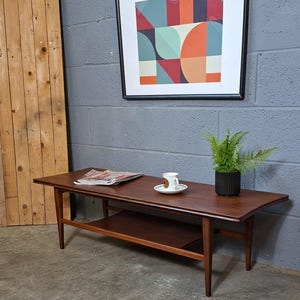 May include: A rectangular wooden coffee table with a lower shelf and tapered legs. On the table sits a magazine, a white ceramic cup and saucer with a colourful pattern, and a green plant in a black ribbed pot. A framed geometric print hangs above.