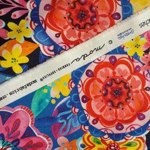 May include: A vibrant fabric with a floral pattern in shades of red, orange, pink, yellow, and blue. The fabric features large, stylized flowers and leaves against a dark background. A white strip with text is diagonally across the fabric.