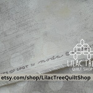 May include: A close-up of a white fabric with a gray geometric pattern. The fabric is labeled "Moda Fabrics" and "Lilac Tree Quilt Shop" with the website address etsy.com/shop/LilacTreeQuiltShop.