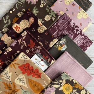 May include: A collection of folded fabric squares featuring various autumn-themed prints including fall leaves, pumpkins, sunflowers, and tea-related motifs in shades of brown, tan, plum, and cream, with a Moda Windfall fabric label visible.