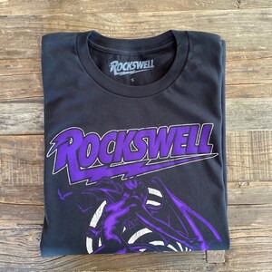 May include: Black t-shirt with a purple graphic of a winged figure and the text "ROCKSWELL" in a purple, stylized font.