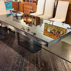 May include: A rectangular dining table featuring a reflective mirrored glass top with a dark metal frame. The table is supported by a central pedestal base with a mirrored finish. The surface reflects the surrounding room, including chairs.