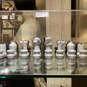 May include: A set of white ceramic chess pieces displayed in a glass case. The pieces are arranged in a chessboard pattern, with the white pieces on the left and the black pieces on the right.