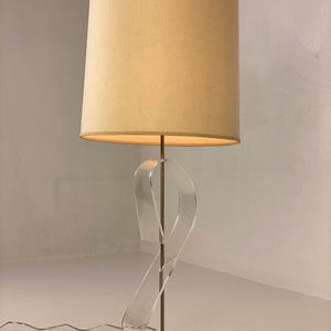 May include: A vintage table lamp with a white fabric lampshade and a clear acrylic base. The base is shaped like a twisted ribbon and has a silver metal base.