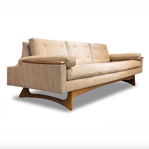 May include: A three-cushion sofa upholstered in a textured beige and multi-colored fabric. The sofa features wooden armrest caps and a curved wooden base frame with arched legs. The back cushions are tufted with buttons in a linear arrangement.