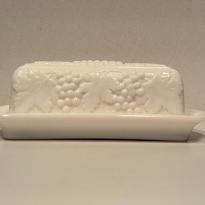 May include: A white, rectangular butter dish with a lid. The lid and base are adorned with a grape and leaf design. The dish is made of a glossy, opaque material.