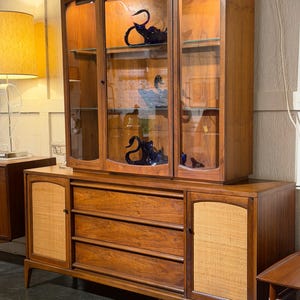 May include: A mid-century modern wooden hutch with a display cabinet and a lower cabinet with drawers. The display cabinet has glass doors and shelves, showcasing blue glass swan figurines. The lower cabinet features woven panels and three drawers.