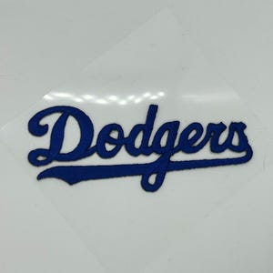 May include: A blue embroidered patch featuring the word "Dodgers" in a stylized font. The patch is in a cursive style and is likely for application on clothing or accessories. The patch is on a clear backing.
