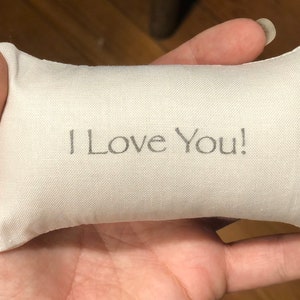 Puede incluir: Una peque&ntilde;a almohada blanca con las palabras "&iexcl;Te amo!" bordadas.