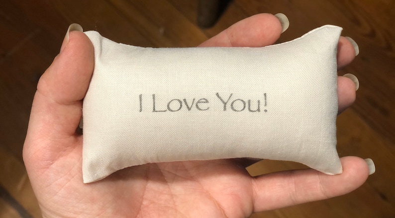 Puede incluir: Una peque&ntilde;a almohada blanca con las palabras "&iexcl;Te amo!" bordadas.
