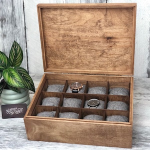 May include: Wooden watch box with a hinged lid, featuring compartments with gray cushions for watch storage. The lid is engraved with a message. A potted plant is on the left. The box is made of light brown wood.