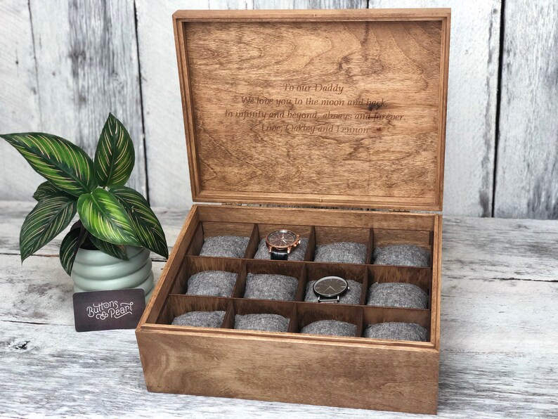 May include: Wooden watch box with a hinged lid, featuring compartments with gray cushions for watch storage. The lid is engraved with a message. A potted plant is on the left. The box is made of light brown wood.